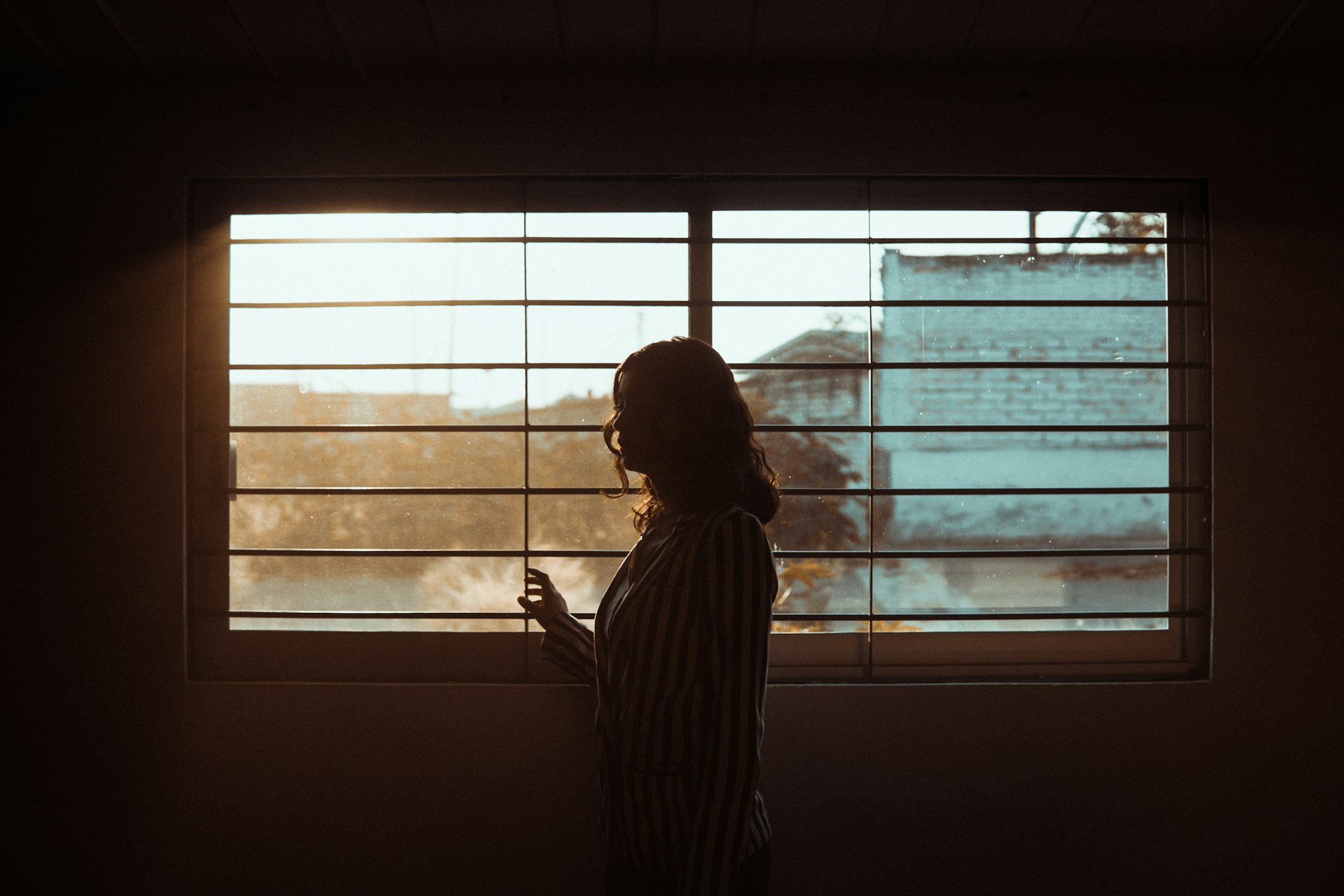 Vrouw staat als silhouet voor een raam met jaloezieën in warm licht