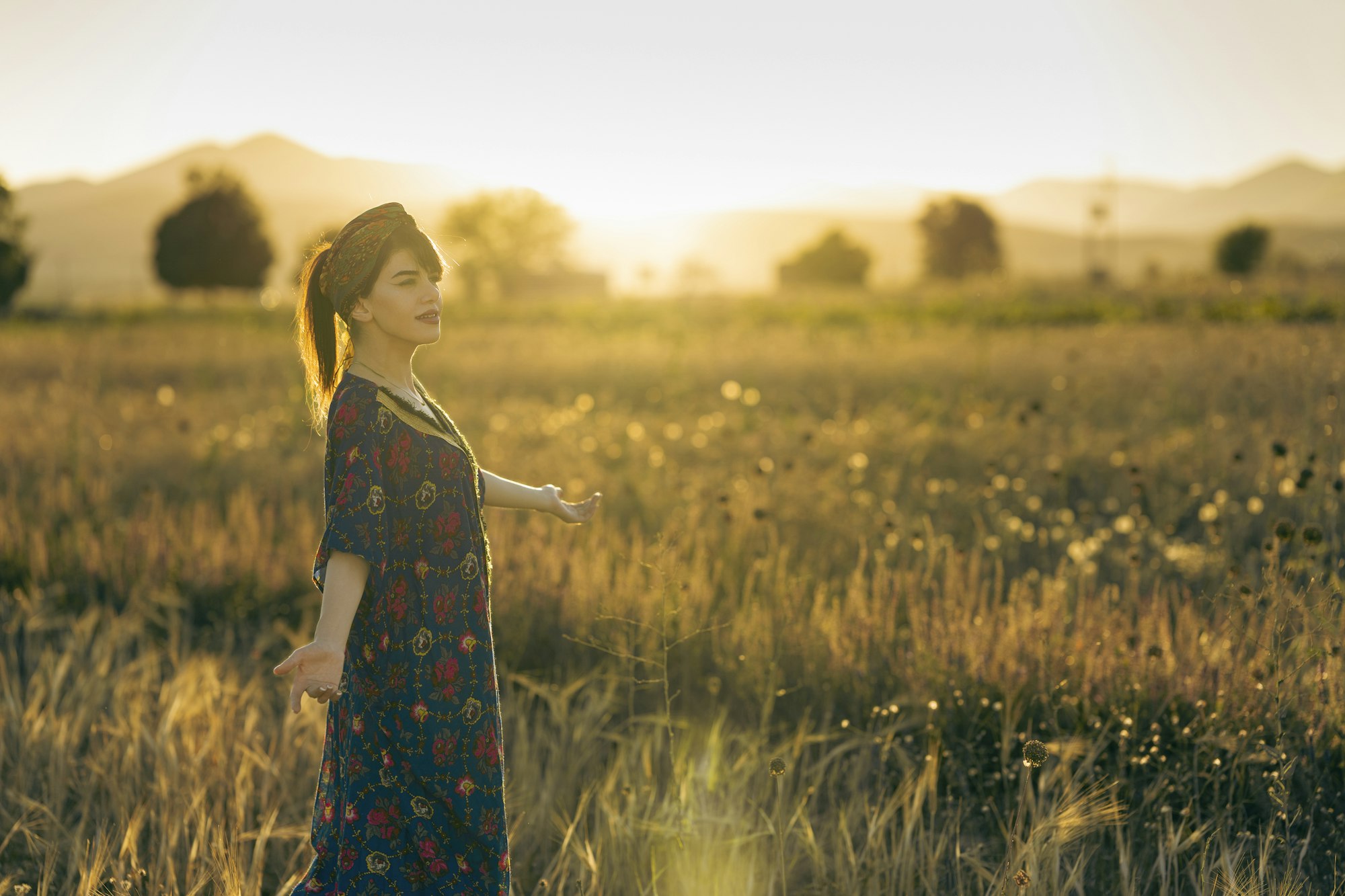Vrouw in bloemenjurk loopt met open arm door een veld bij zonsondergang