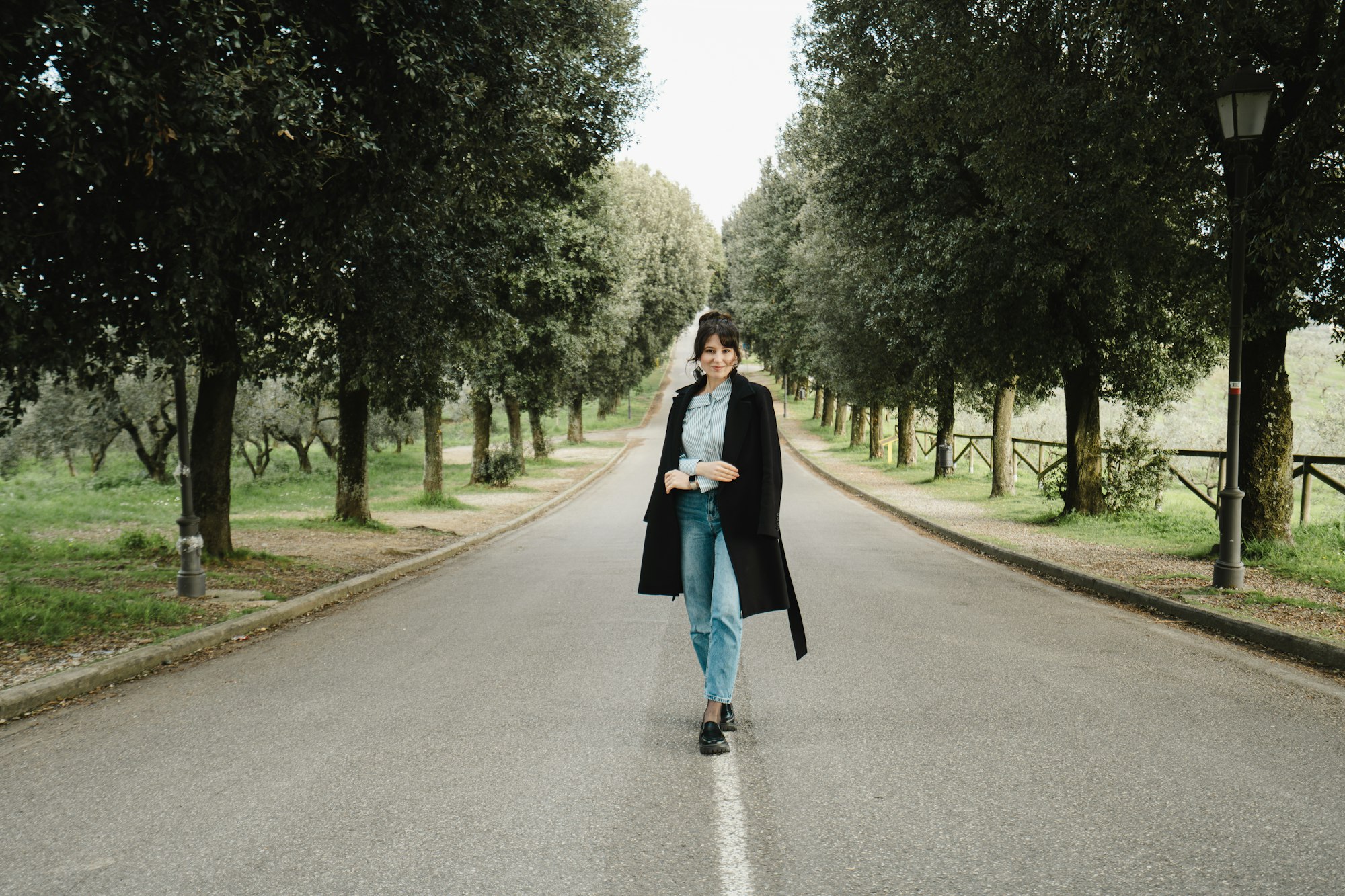 Vrouw loopt in een lange jas midden over een weg tussen hoge bomen