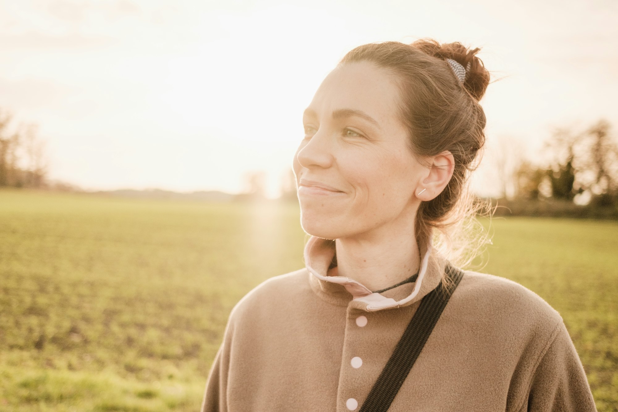 Vrouw in kameelkleurige jas kijkt rustig opzij in avondlicht boven een veld