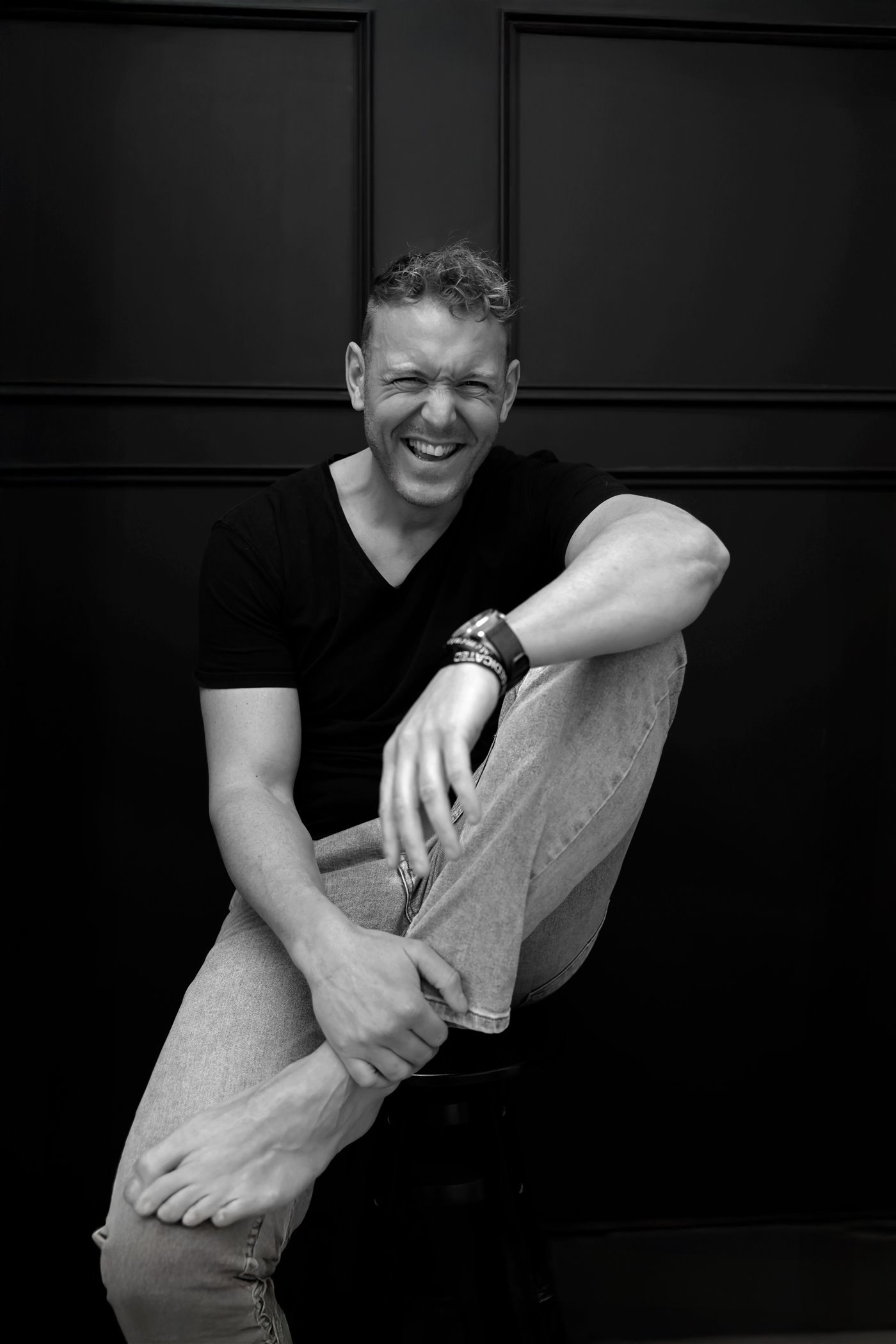 Jonathan Bouter lacht hartelijk, zittend op een kruk met blote voeten